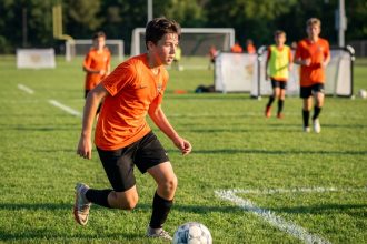 Jeune joueur de football en stage d'été dribblant un ballon sur un terrain gazonné sous la supervision d'un formateur expérimenté